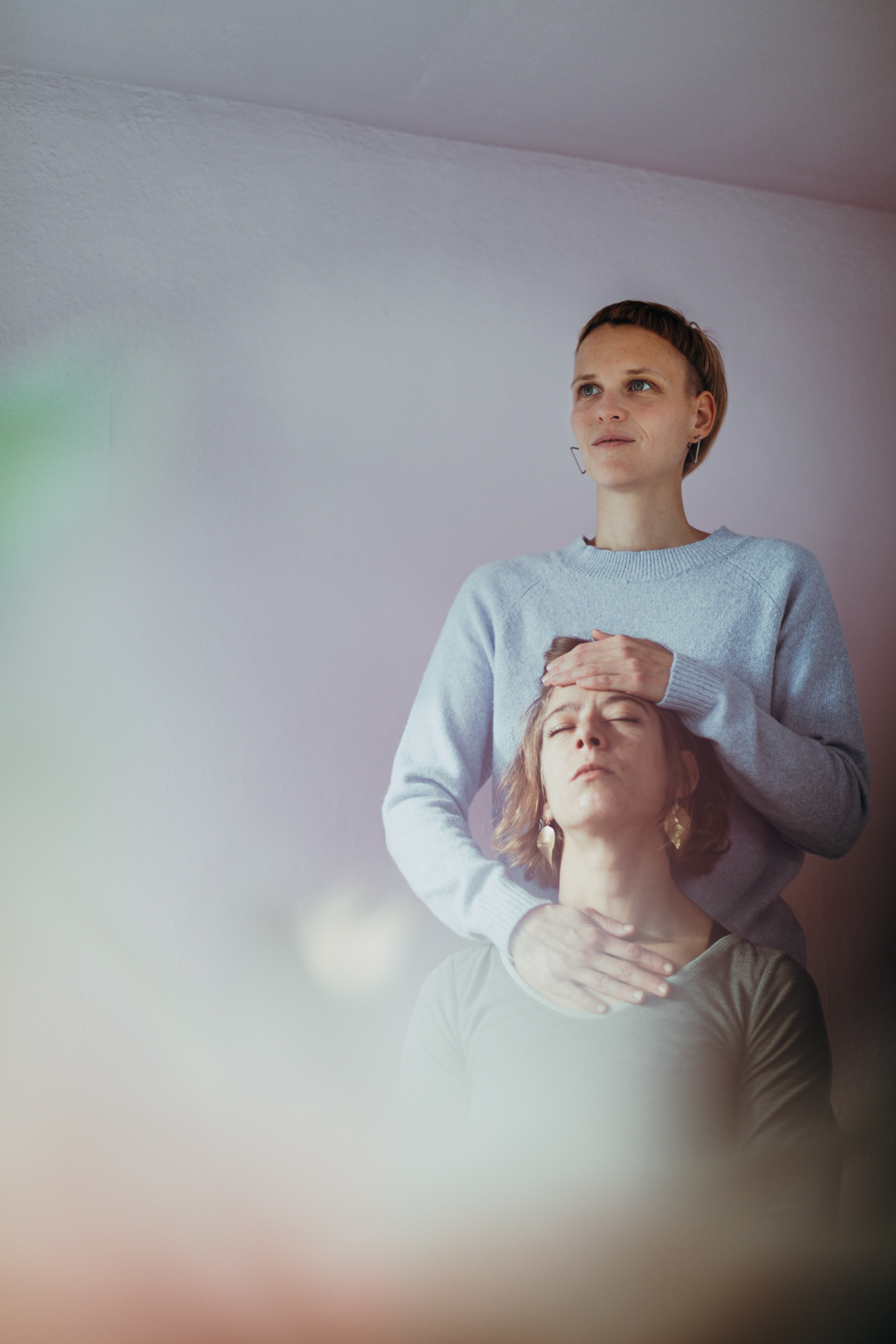 Manu steht hinter einen anderen Person. Die Person ist entspannt. Sie legt ihren Kopf an Manus Oberkörper. Manu legt eine Hand auf die Szirn der Person. Die andere Hand legt Manu auf die Brust der Person.