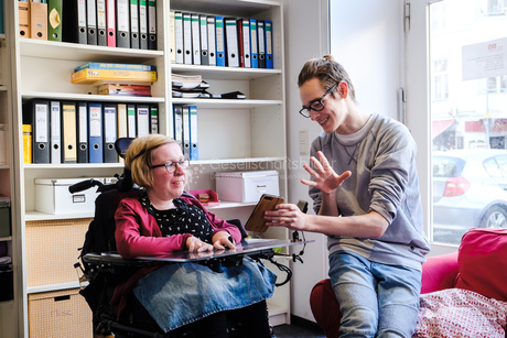 Zwei Personen sind in einem gemütlichen Raum. es gibt ein Sofa und ein Bücherregal. Eine Person sitzt im Rollstuhl. Sie hört der anderen Person zu. Die andere Person zeigt etwas auf ihrem Telefon.