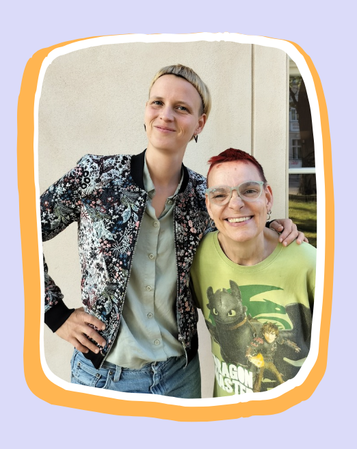 Manu und Tine stehen Arm in Arm nebeneinander. Tine lacht breit. Tine hat kurze rote Haare und ein grünes T-Shirt an. Manu löchelt, hat kurze blonde Haare und eine blumige Jacke an.