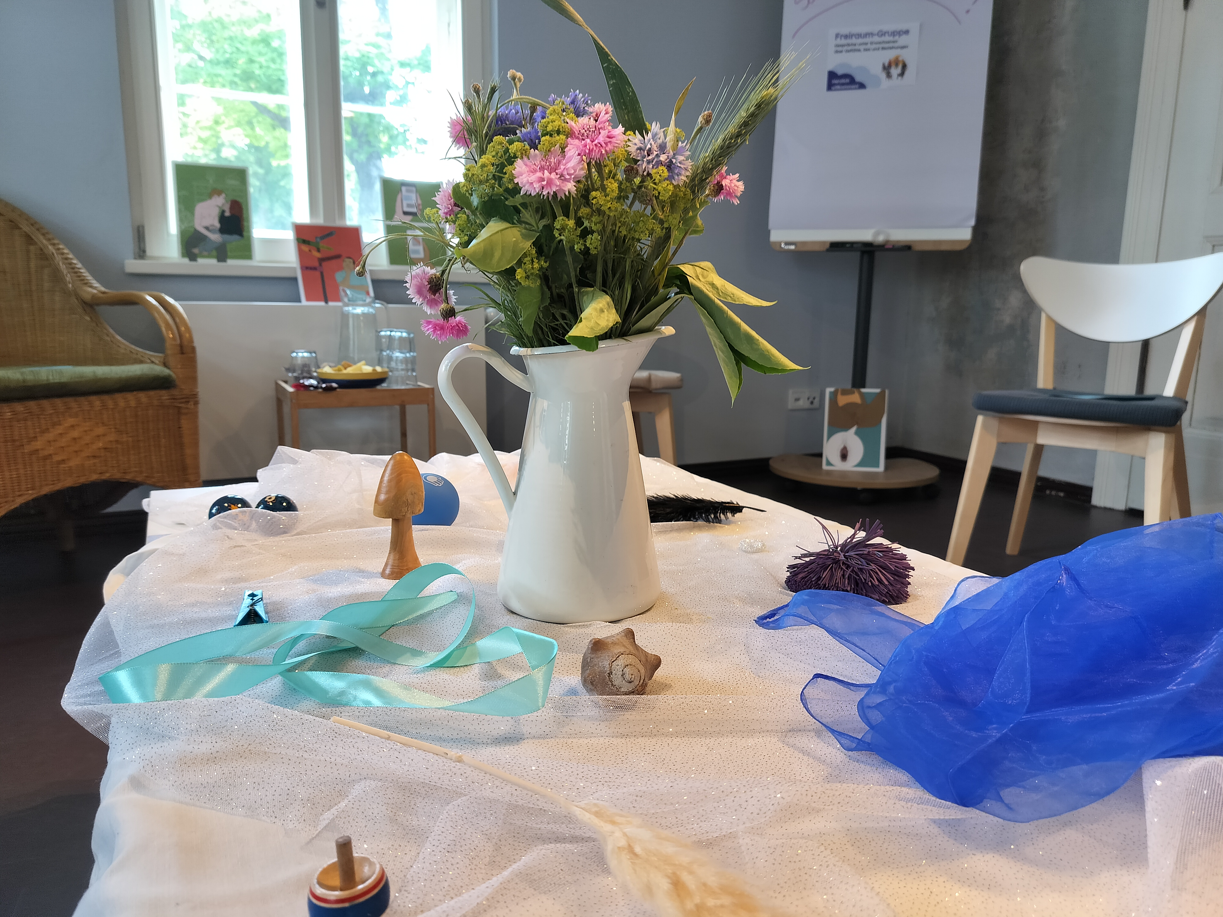 Auf einem Tisch steht eine Blumenvase. Ringsherum liegen verschiedene Gegenstände_ Ein Holzpilz, eine Muschel, ein Band, ein Tuch. Im Hintergrund sind Bilder. Die Bilder sind gezeichnet. Menschen küssen sich auf den Bildern.