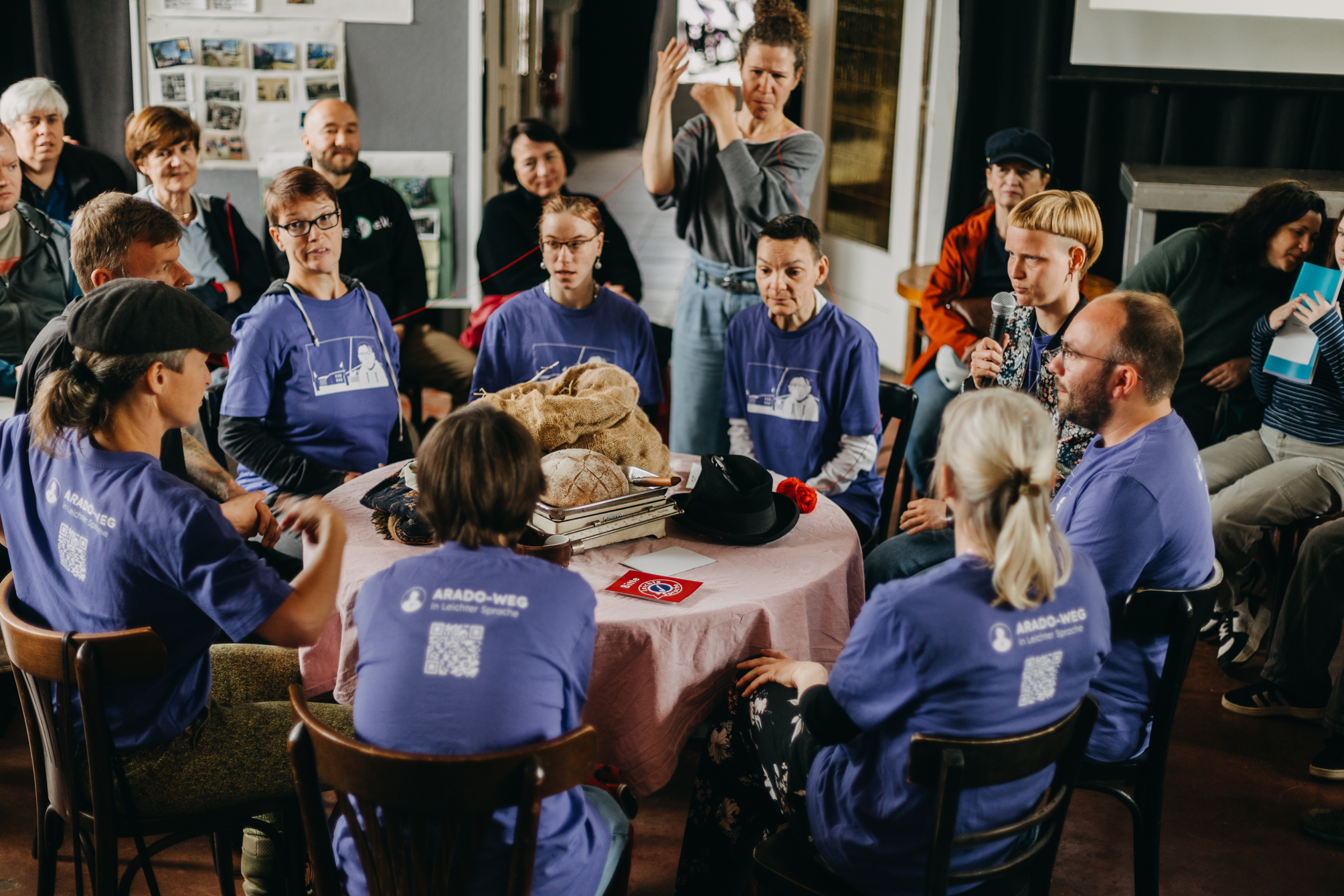 9 Leute sitzen um einen Tisch herum. Die meisten tragen ein lila T-Shirt. Alle schaue konzentroert. Auf dem Tisch in der Mitte liegen verschiedene Gegenstände. Im Hintergrund ist eine DGS-Dolmetscherin. Sie hat ein rotes Band über den Schultrern. Rindsherum sitzen Menschen in kreisen und schauen zu.