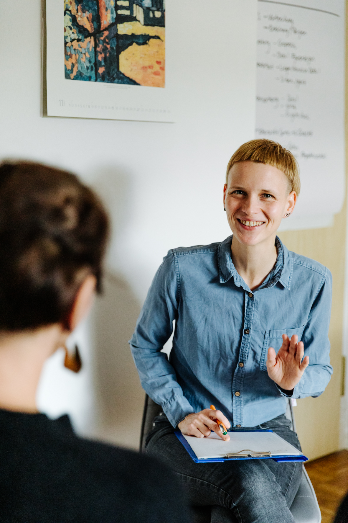 Manu sitzt einer Fachkraft gegenüber, lächelt und erklärt etwas. Die Fachkraft ist von hinten zu erkennen, An der Wand hängt ein Kalender und ein Flipchart.