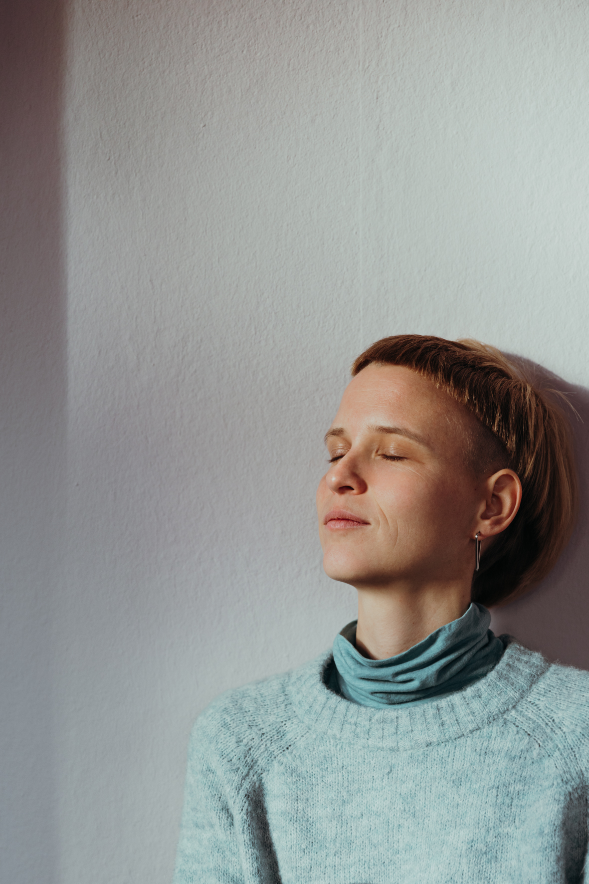 Manu lehnt an einer Wand. Sie hält ihr Gesicht in die Sonne. Sie hat die Augen geschlossen. 